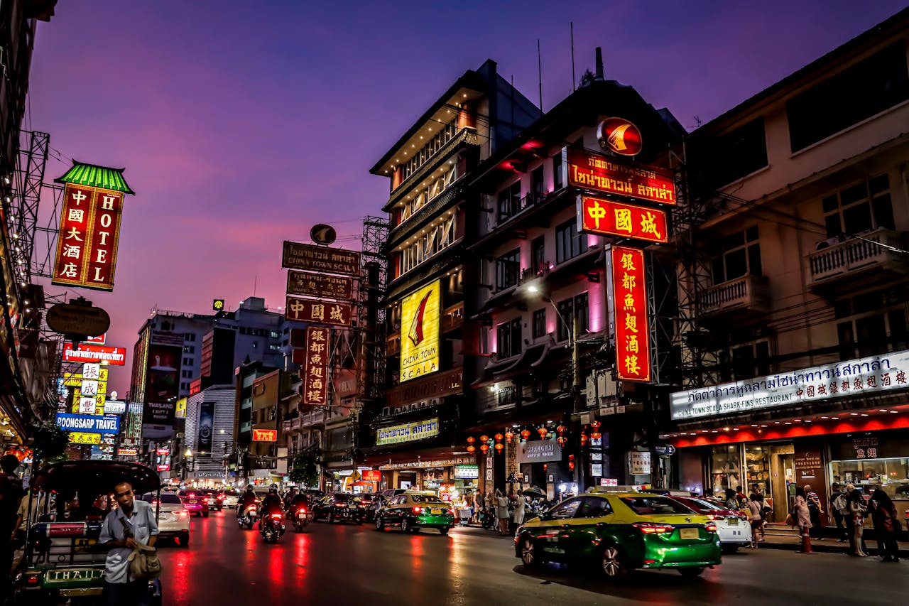 Colorful illuminated signs light up Bangkok's Chinatown street at dusk, showcasing urban life and culture.