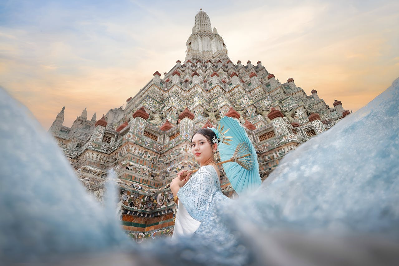 Woman in traditional attire with parasol in front of Wat Arun temple at sunset in Bangkok, Thailand.